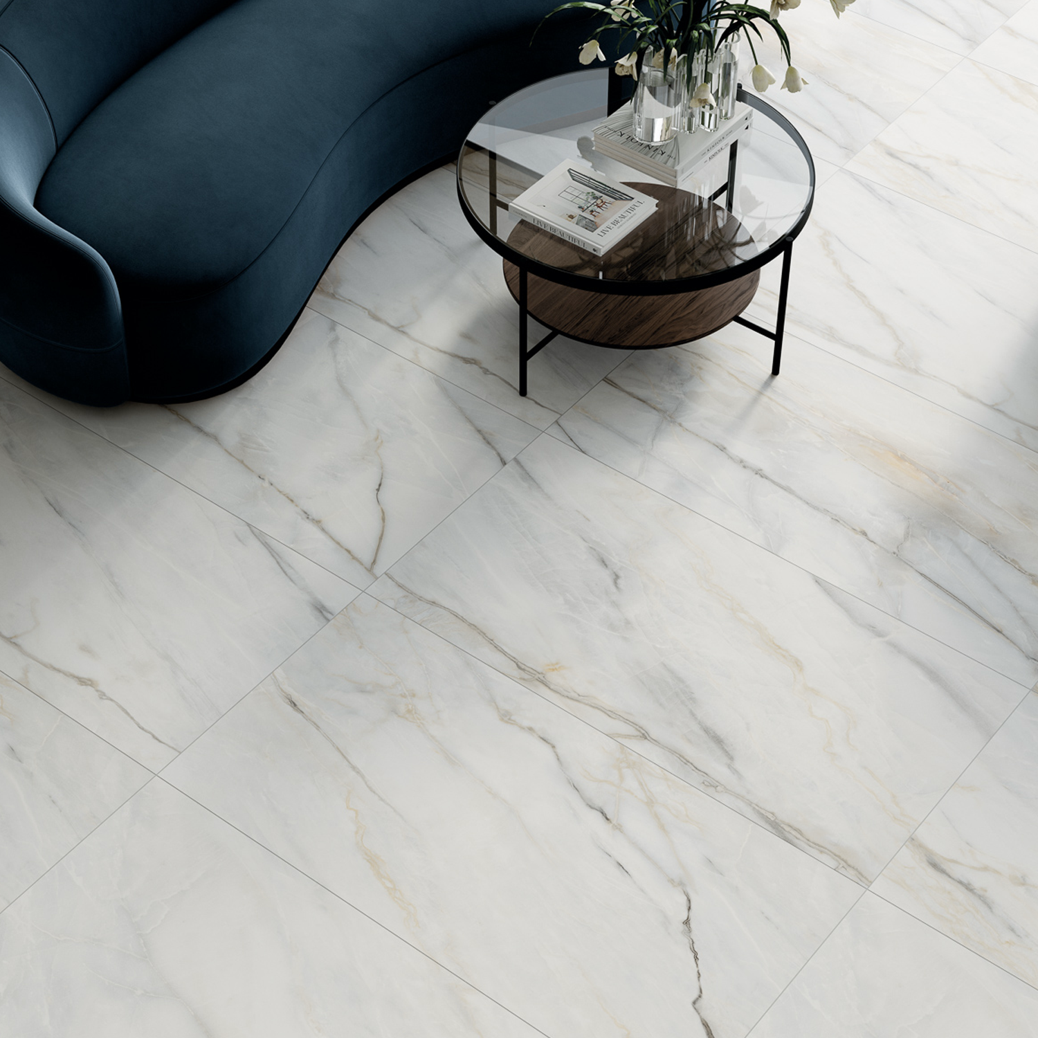 Curved dark blue sofa with a round glass coffee table on Classic Boutique - Calacatta Bluette white marble floor tiles featuring grey and beige veining. A plant, a magazine, and a cup sit on the table. Modern and minimalist living room decor.