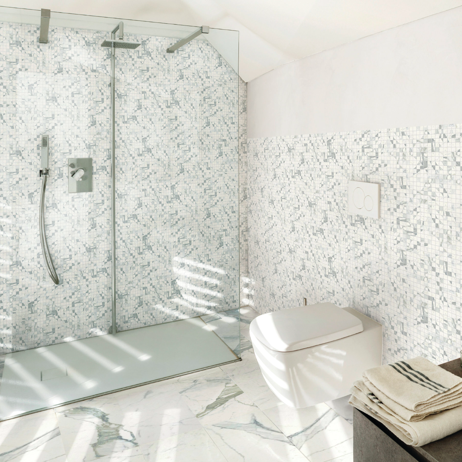 Modern bathroom featuring a walk-in glass shower, mosaic tile walls, wall-mounted toilet, and neatly folded towels atop a dark counter. Sunlight streams in, casting shadows across the Classic - Statuarietto marble floor.
