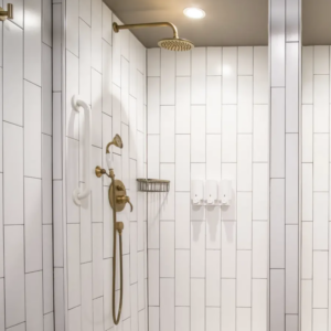 Modern shower with Color Collection - MG White Ice vertical tiles, gold fixtures including a rainfall showerhead and handheld sprayer, a white grab bar, a wire basket, and three white wall-mounted soap dispensers. Ceiling light above.