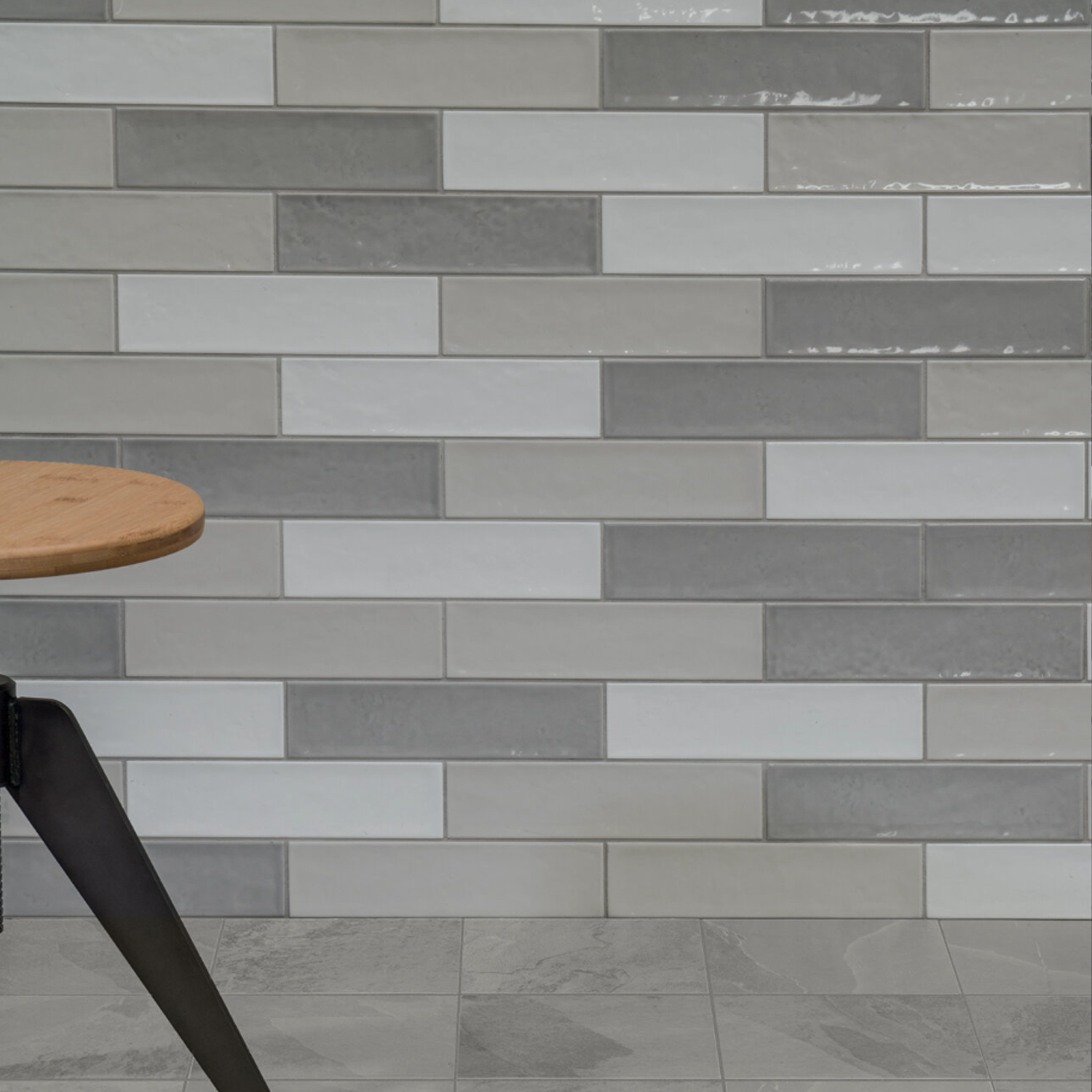 A partial view of a wooden table with black legs is in front of a wall featuring Cromia26 - Grigio 1 staggered gray and white rectangular tiles, set above a gray tiled floor.