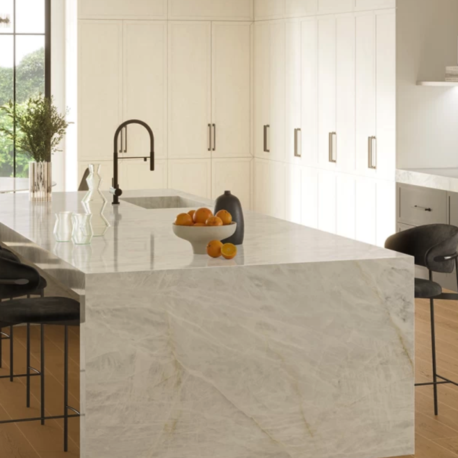 Modern kitchen featuring a large Crystal White Bookmatch marble island, complemented by a black faucet, black stools, and a bowl of oranges. Light wood floors and tall white cabinets line the background as sunlight streams through the window.
