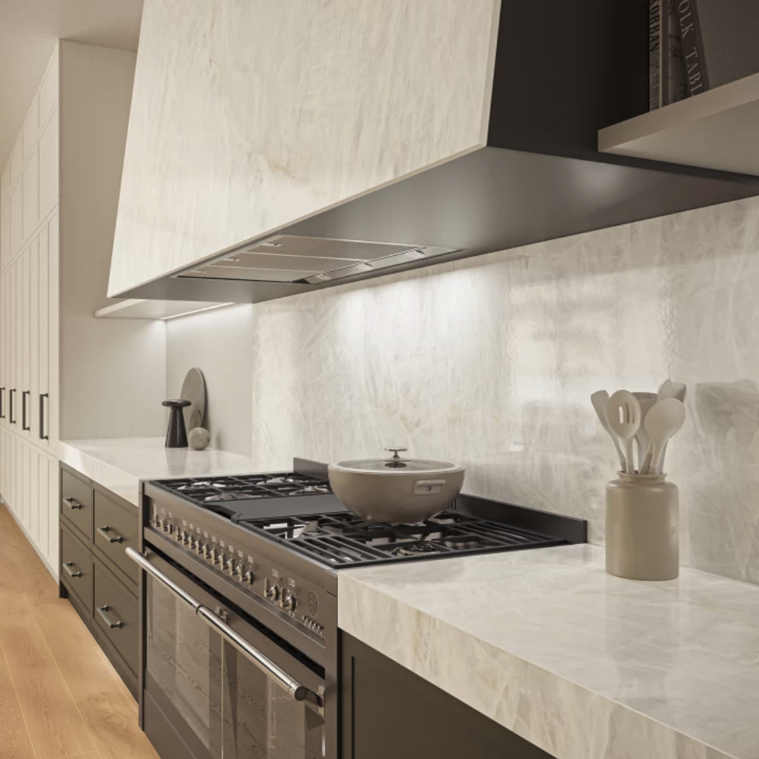 A modern kitchen featuring Crystal White Bookmatch countertops and backsplash, a black oven range with a gray pot, white utensils in a holder, and sleek cabinetry. Warm wooden flooring and under-cabinet lighting highlight the clean, elegant design.