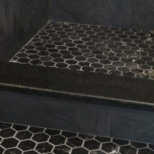 A shower featuring Curbs - Black hexagonal floor tiles with black grout, adjacent to a step or ledge; outside the shower, matching Curbs - Black tiles are set off by white grout lines.