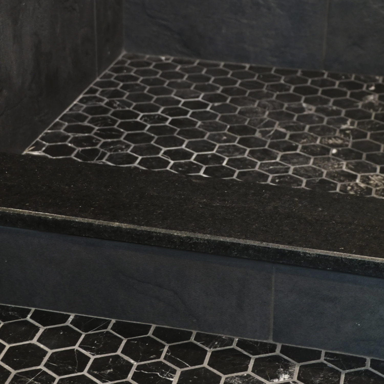 A shower featuring Curbs - Black hexagonal floor tiles with black grout, adjacent to a step or ledge; outside the shower, matching Curbs - Black tiles are set off by white grout lines.