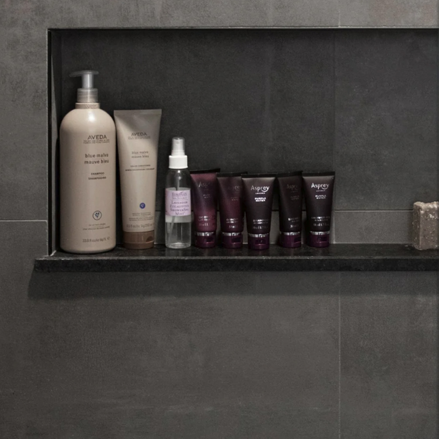 A Curbs - Black shower niche with six Aveda hair and skincare products, a spray bottle, and a bar of soap on a gray tiled wall.