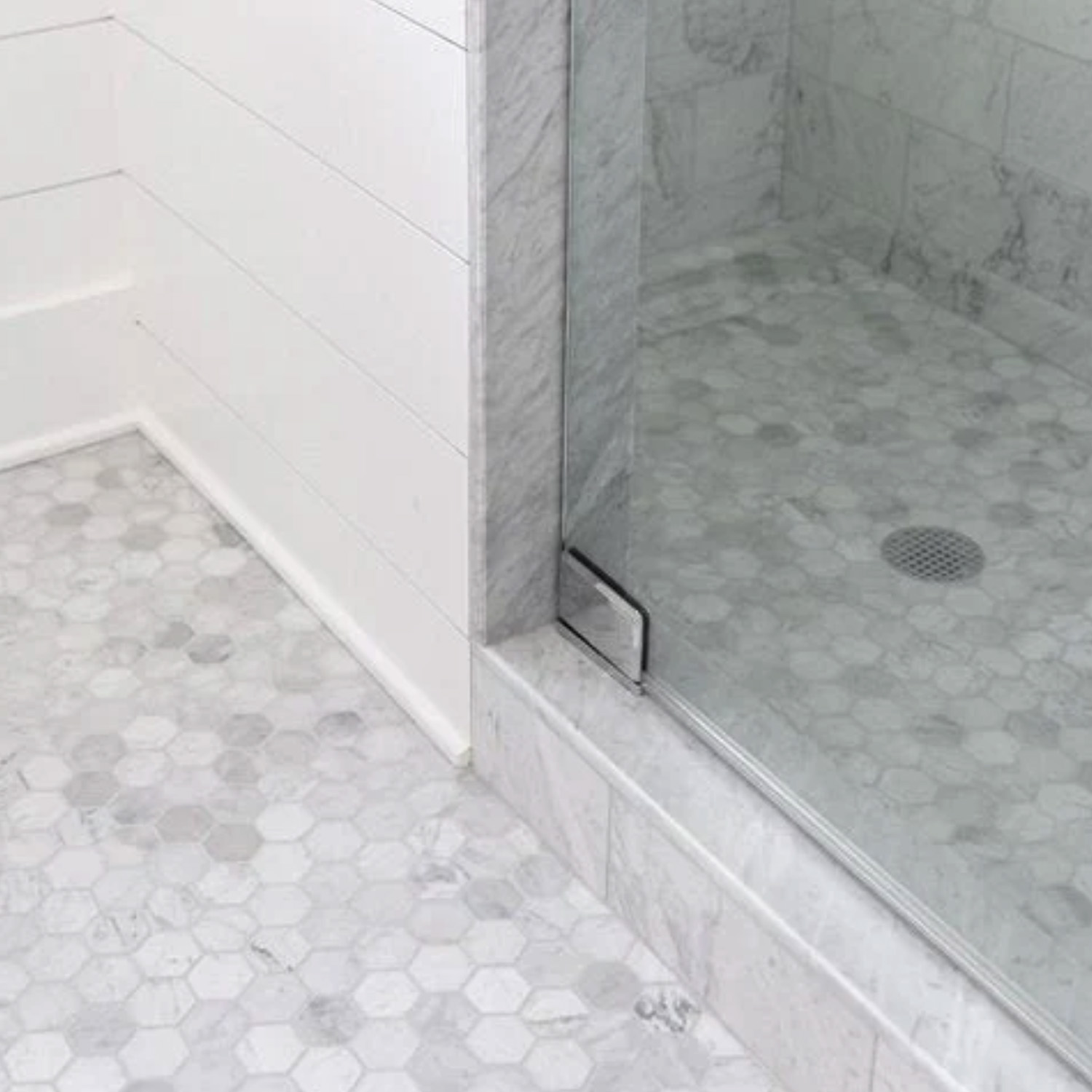 A bathroom corner with a glass shower enclosure features light gray and white hexagon floor tiles, while the shower curb is finished with Curbs - Venatino Carrara and the walls are completed with marble tile and white paneling.