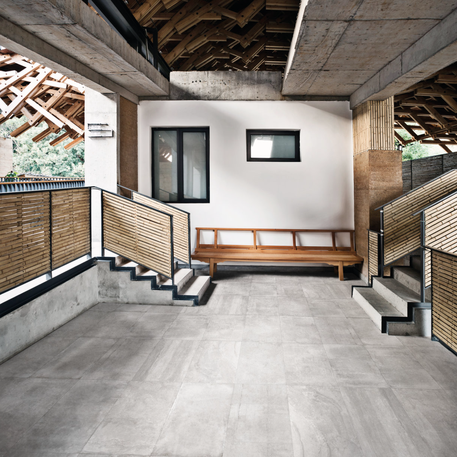 Open indoor area with Ecocrete - Aqua gray tiled floor, a wooden bench against a white wall, two staircases featuring bamboo railings, and unfinished wooden beams above. Natural light streams in from the openings.