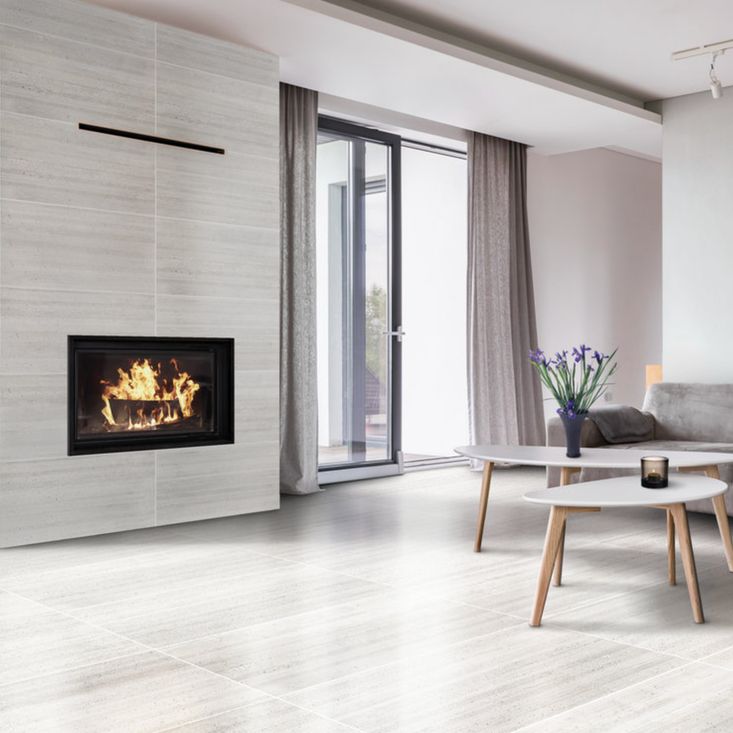 Contemporary living room featuring Ecocrete - Melted Ice light wood floors, a cozy lit fireplace, two round tables, a gray sofa, vibrant purple flowers in a vase, and expansive glass doors adorned with curtains.
