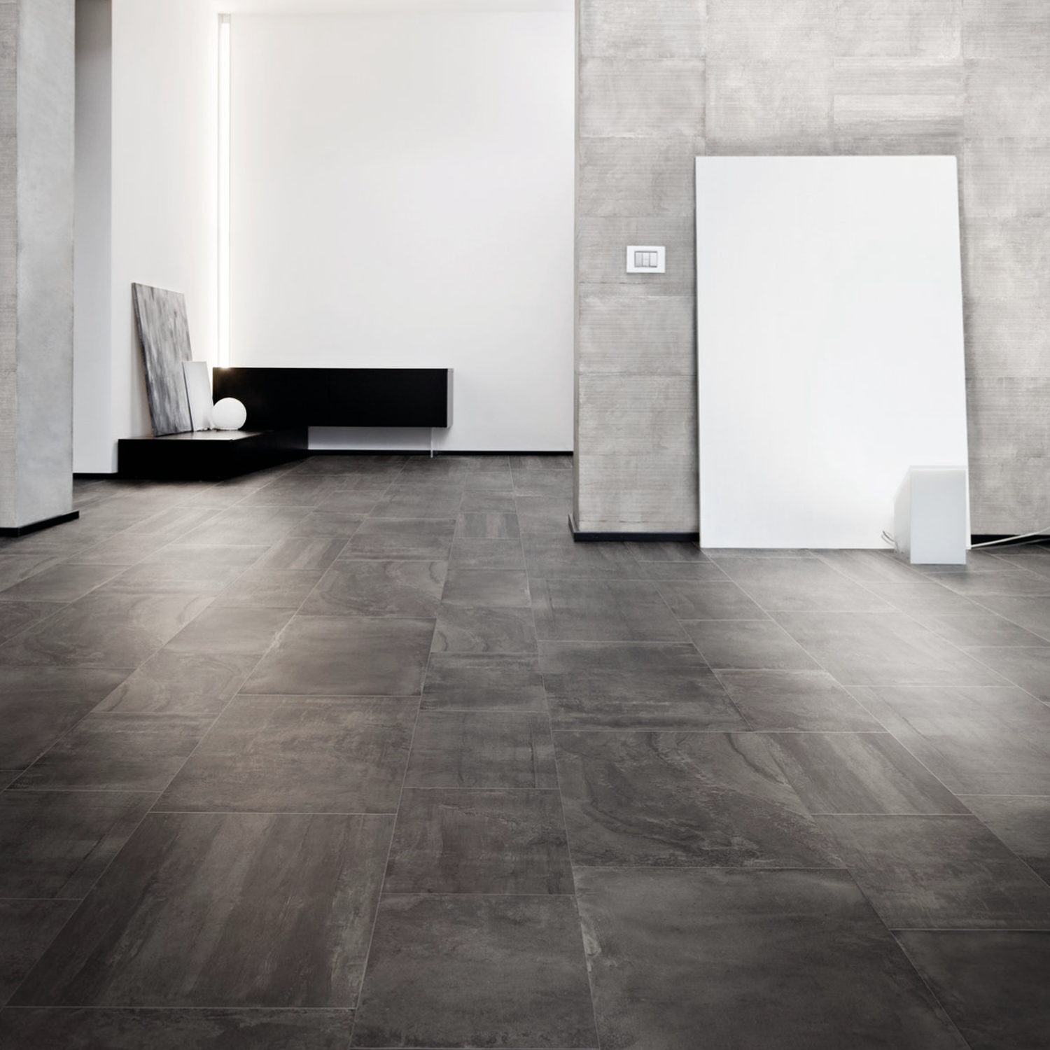 Minimalist room with large Ecocrete - Weathered Black floor tiles, light gray textured walls, a low black bench, a white spherical object, and two large canvases leaning against the wall. Bright vertical light accent in the back corner.