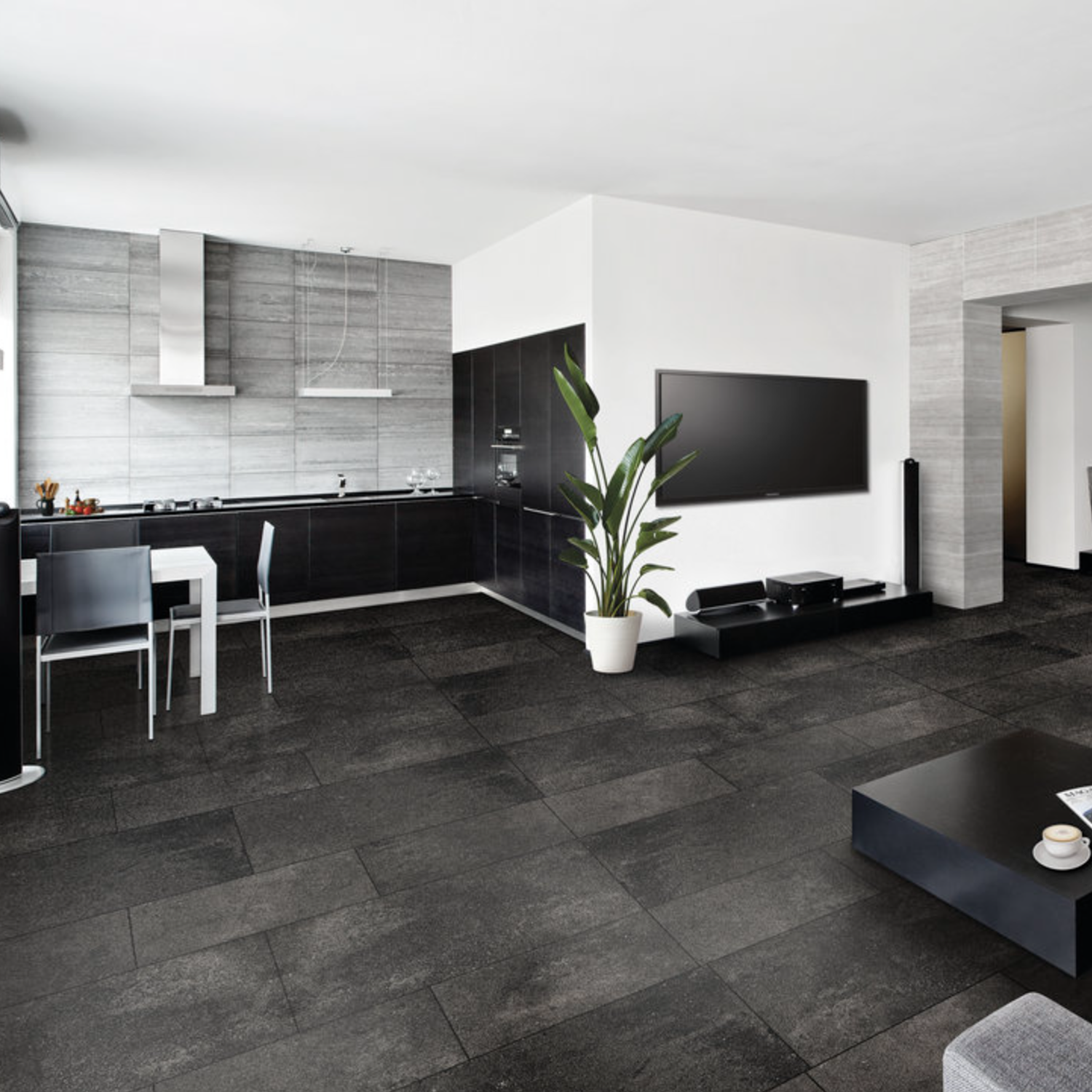 Modern open-plan kitchen and living room featuring Ecocrete - Weathered Black cabinets, a wall-mounted TV, dining table, potted plant, and dark tile flooring. The space is adorned with minimalist decor and a neutral color palette throughout.