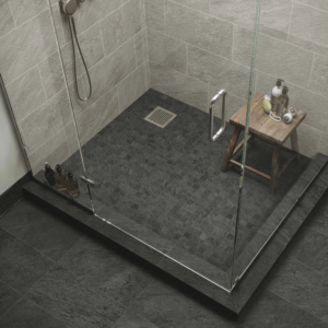 Modern glass shower featuring Element - Carbon dark stone floor tiles, beige wall tiles, and a small wooden stool holding soap and a sponge. Bottles of toiletries sit on the floor beside the glass enclosure.