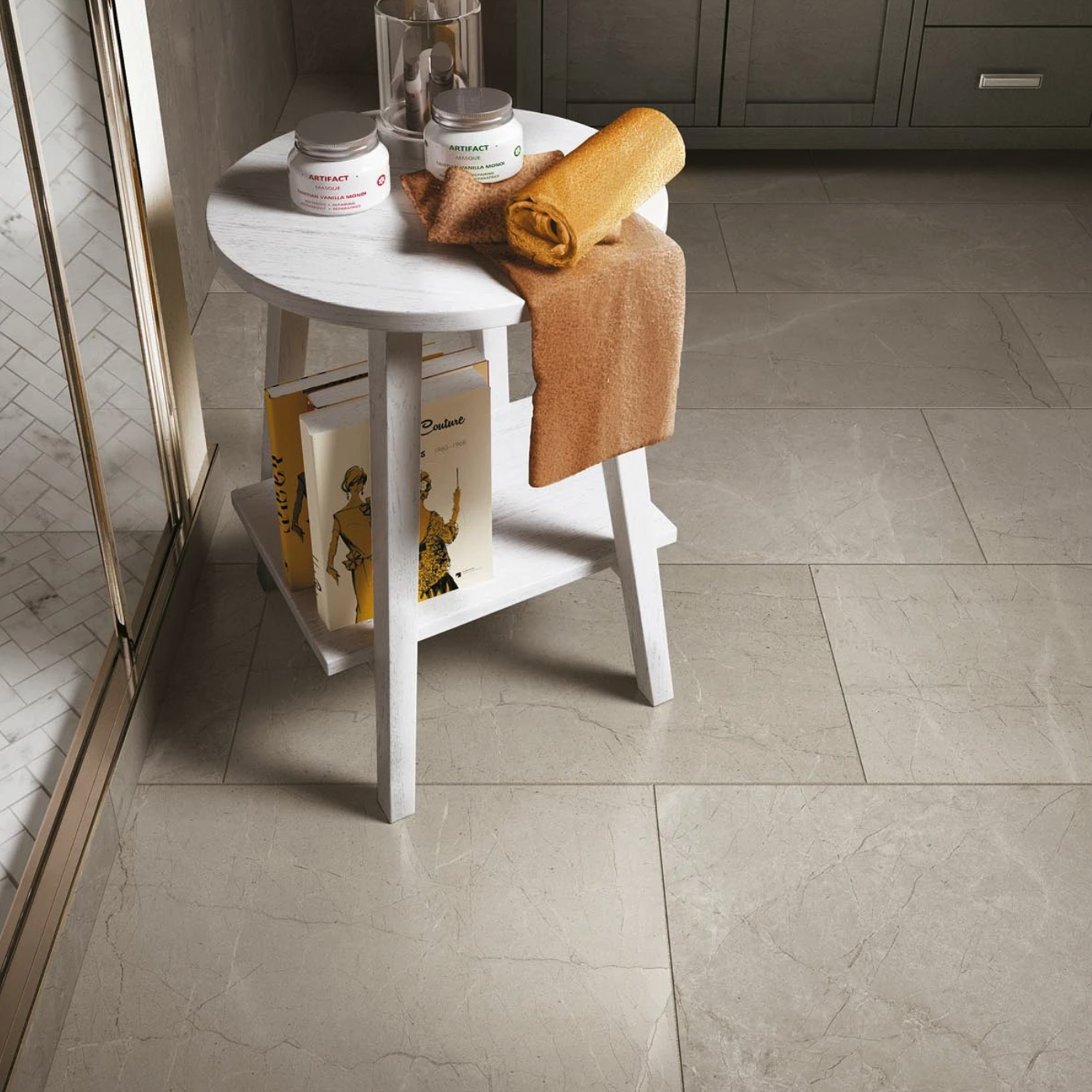 A small white table sits on an Eon - Corinthian Gray tiled bathroom floor, holding two jars, a glass, an orange towel, and books with yellow covers next to a glass shower.