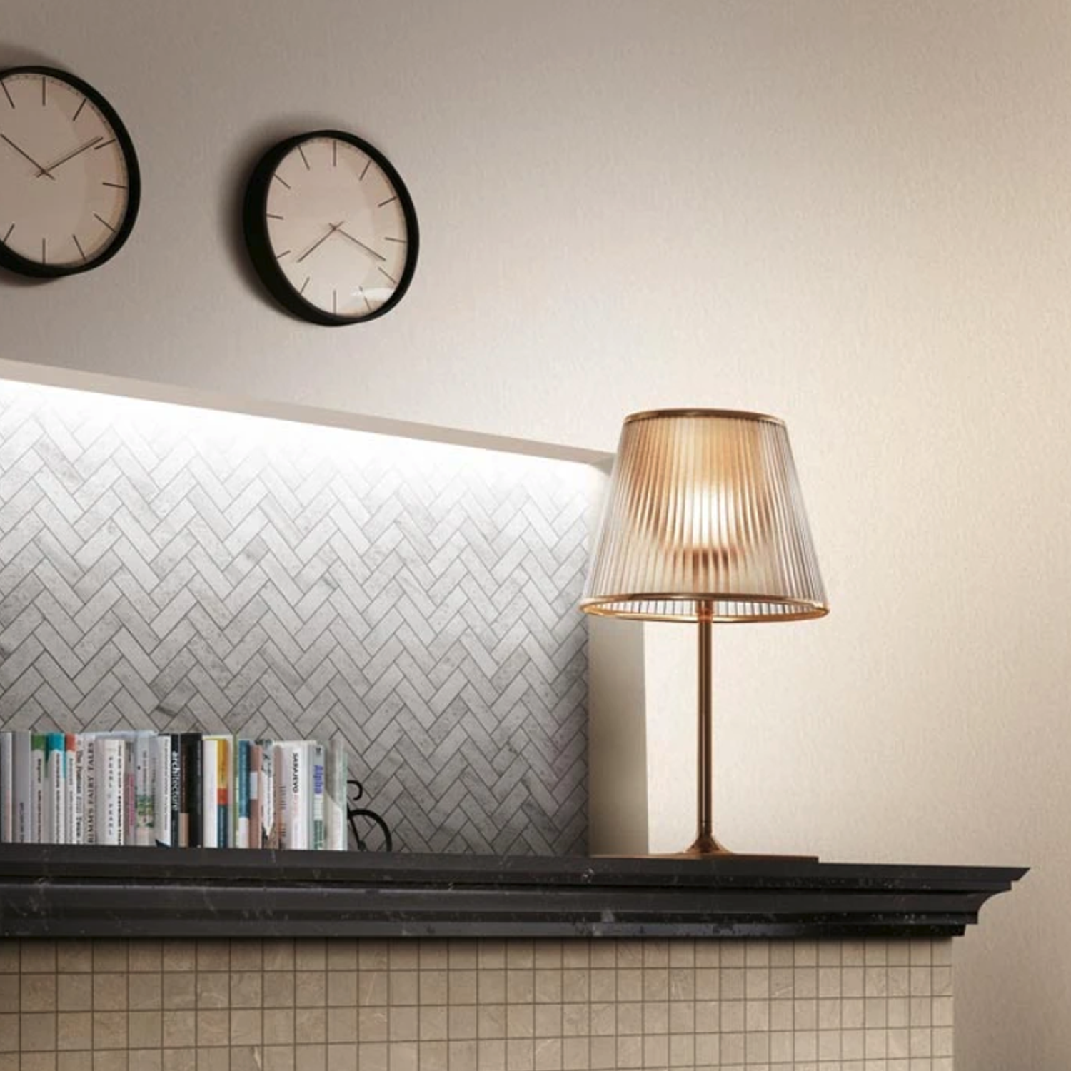 A modern interior features the Eon - Eldorado table lamp on a tiled shelf, books neatly arranged, and two round wall clocks above a herringbone-patterned backsplash.