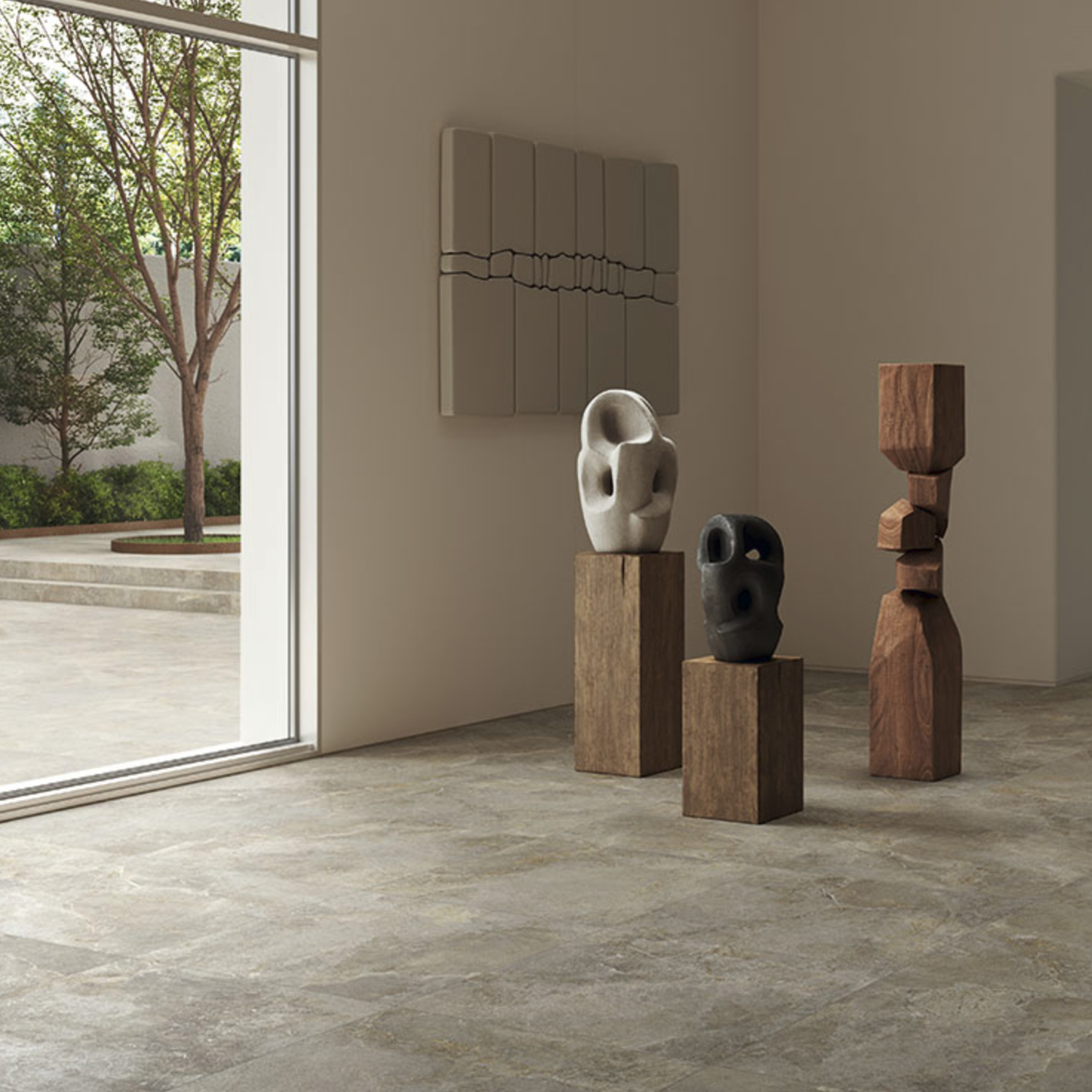 A minimalist room featuring three abstract sculptures displayed on wooden pedestals near a large window adorned with Everstone - Ever Brown. Sunlight streams in, illuminating the artworks, while a tree and patio are visible outside.