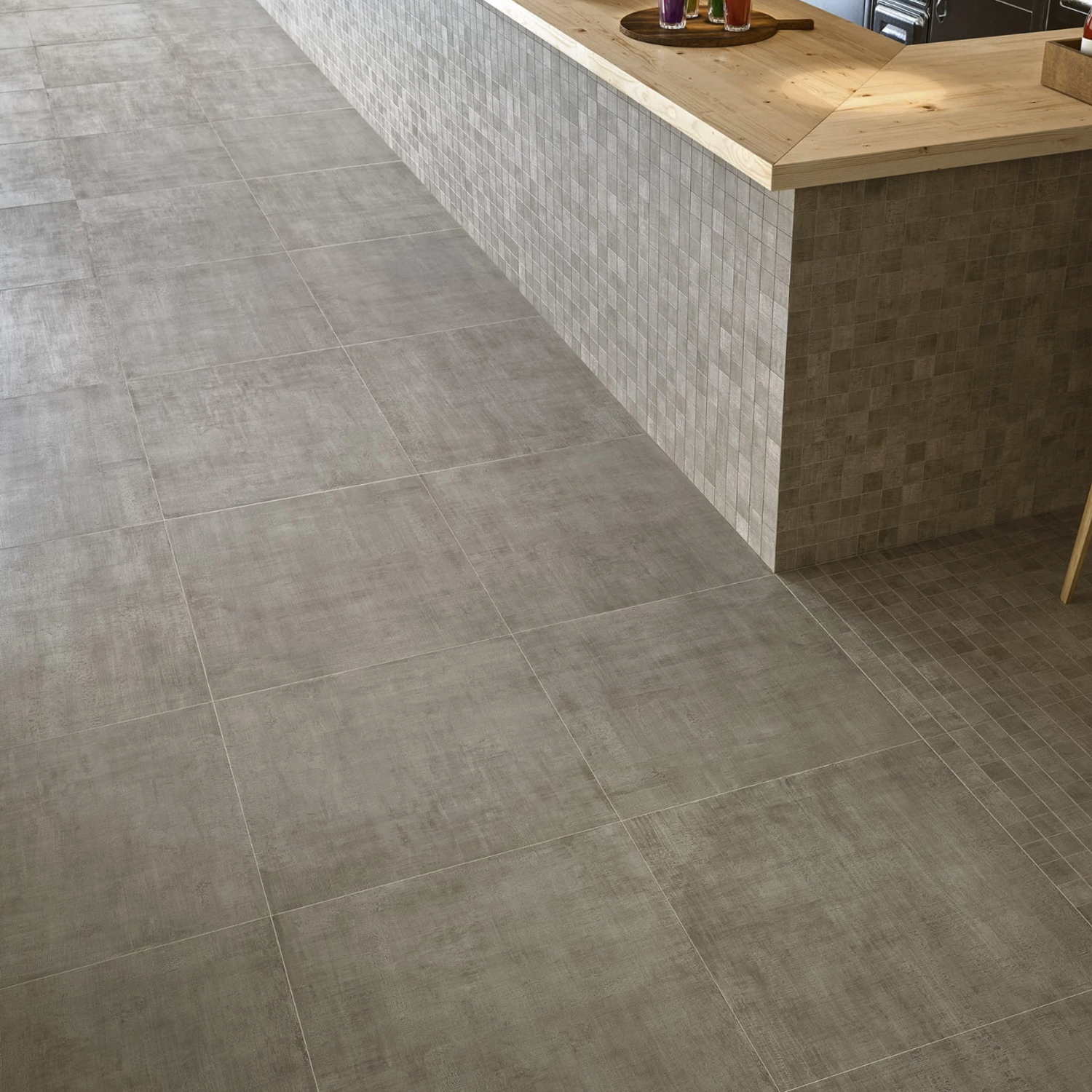 A modern kitchen featuring a wooden countertop and light-colored tiled surfaces, with large Fray - Gray tiles on the floor. The counter base is covered in small beige tiles, and various kitchen items are visible on the countertop.