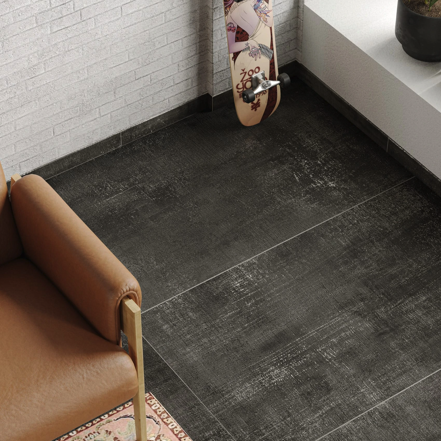 A Fray - Metal Black chair sits on a large, dark tile floor next to a wall with white brick. A skateboard with colorful graphics leans against the wall. A small section of a patterned rug is visible in the corner.