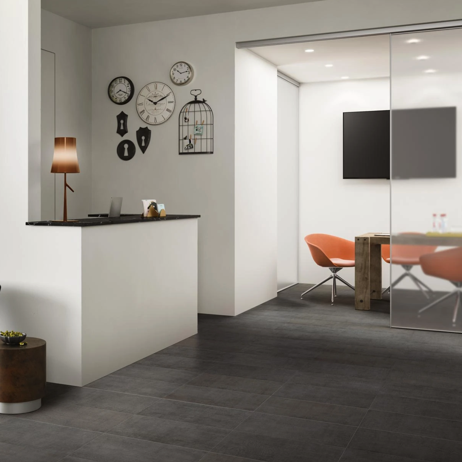 A modern office featuring the Fray - Smoke small reception desk, wall clocks, and a meeting room with a wooden table and four orange chairs situated behind glass doors. The minimalist decor showcases black and white tones.