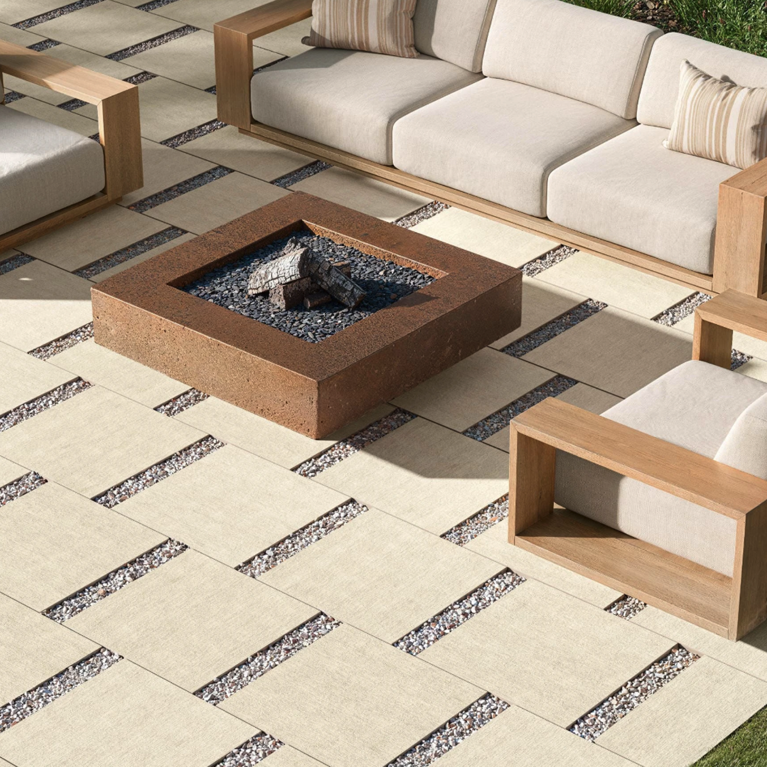 Patio features Fray X - Almond cushioned sofas, wooden armchairs, a square stone fire pit with black rocks, and large light tiles set with gravel-filled gaps. A border of green grass surrounds the patio area.