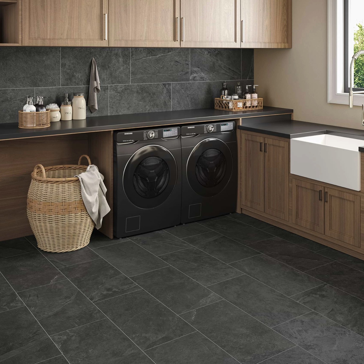 Modern laundry room featuring a Hero - Coal black tile floor, wooden cabinets, a black washer and dryer, a wicker laundry basket, a white farmhouse sink, and various containers on the countertop set against a dark tiled wall.