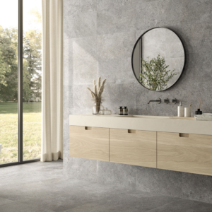 Modern bathroom featuring Ikonite - Grey stone walls, a round mirror reflecting greenery, light wood cabinets, minimalist decor, and a large window showcasing views of trees and grass outside.