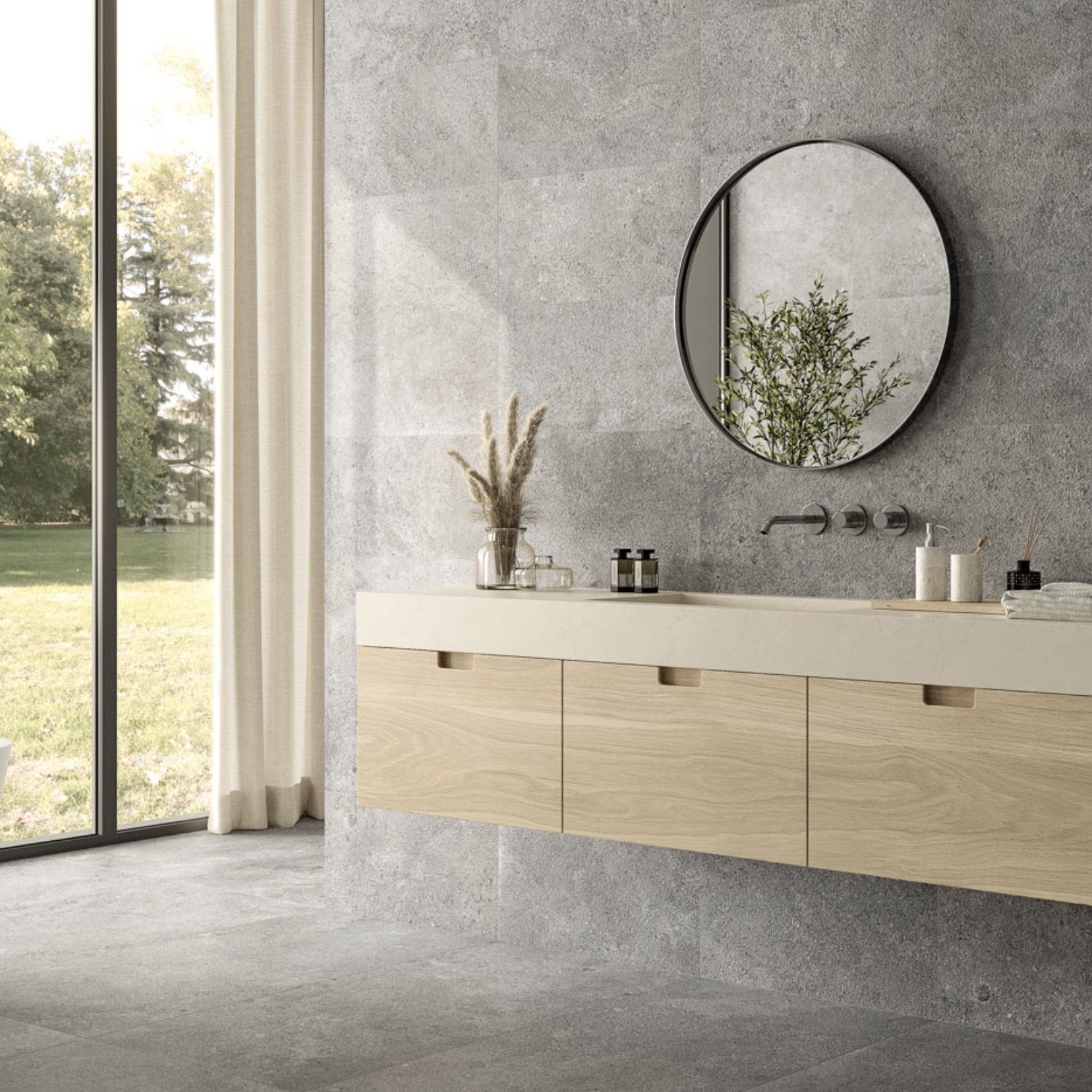 Modern bathroom featuring Ikonite - Grey stone walls, a round mirror reflecting greenery, light wood cabinets, minimalist decor, and a large window showcasing views of trees and grass outside.