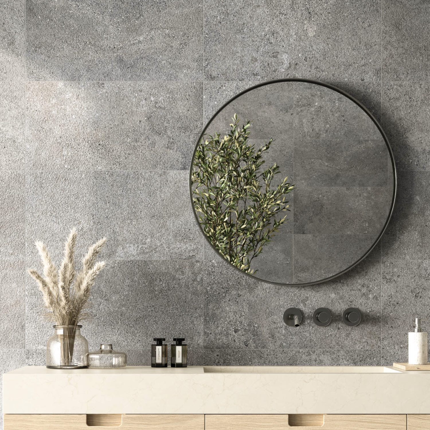 A round Ikonite - Grey mirror hangs on a gray stone wall, reflecting a leafy green branch; below, a beige countertop displays glass jars, vases with dried grass, and soap bottles.