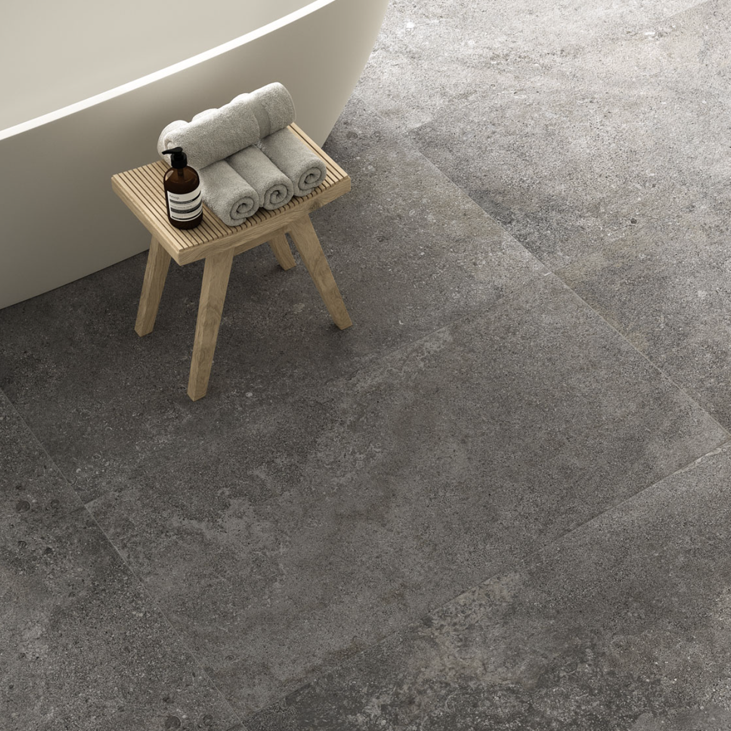A small wooden stool with rolled towels and a soap dispenser sits beside a curved bathtub on an Ikonite - Grey stone tile floor.
