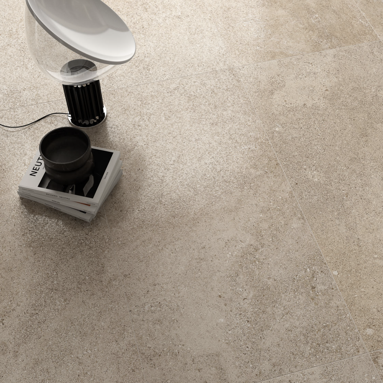 The Ikonite - Walnut lamp, a black cup, and a stack of magazines are arranged on a textured beige tile floor with subtle speckles. The lamp’s cord is partially visible, curving on the floor.