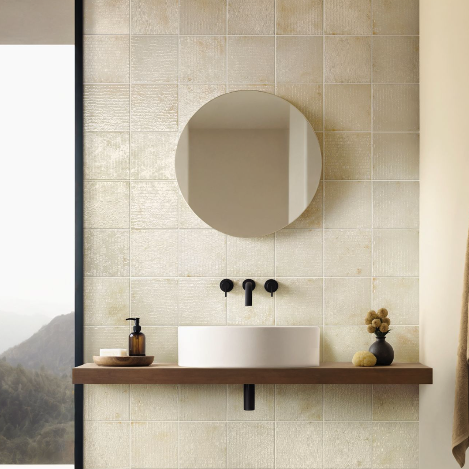 A minimalist bathroom featuring the Inciso - Desert collection, with a round mirror, white vessel sink, wall-mounted black faucet, and a wooden shelf displaying soap and decor. Beige tiled walls serve as the backdrop, while a window frames picturesque mountain views.