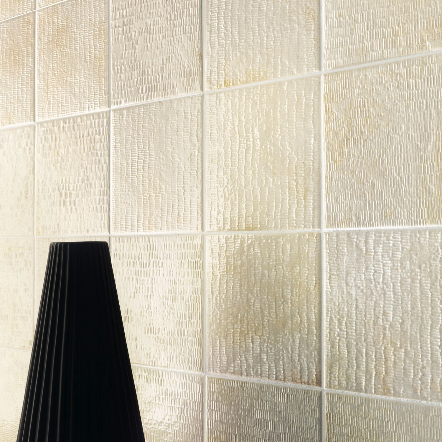 Inciso - Desert tiles, featuring a textured cream color and a subtle wavy pattern, cover the wall. In the bottom left corner, a portion of a black, ribbed lampshade can be seen.