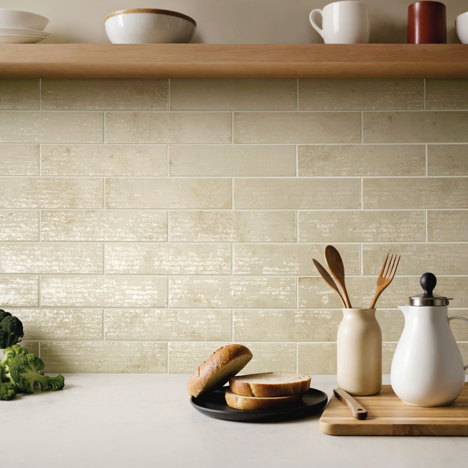 The kitchen counter displays sliced bread on a black plate alongside a jar of wooden utensils and a teapot, with a wooden shelf above holding bowls and cups. The backsplash is tiled in Inciso - Desert, featuring beige rectangular tiles.