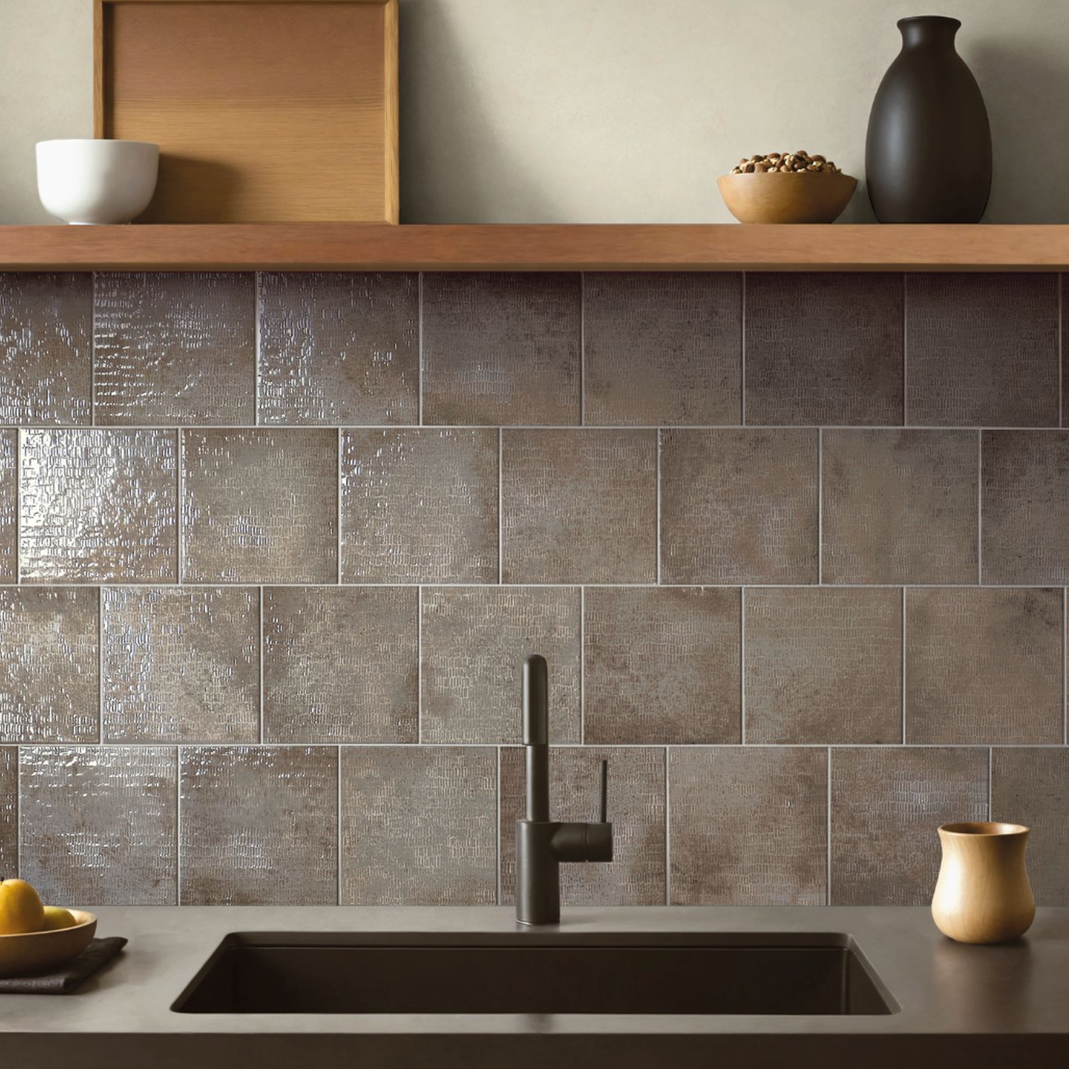 Modern kitchen featuring the Inciso - Pebble matte black sink and faucet, complemented by a brown textured tile backsplash. A wooden shelf displays bowls, a vase, and a tray, while lemons and a small cup rest on the countertop.
