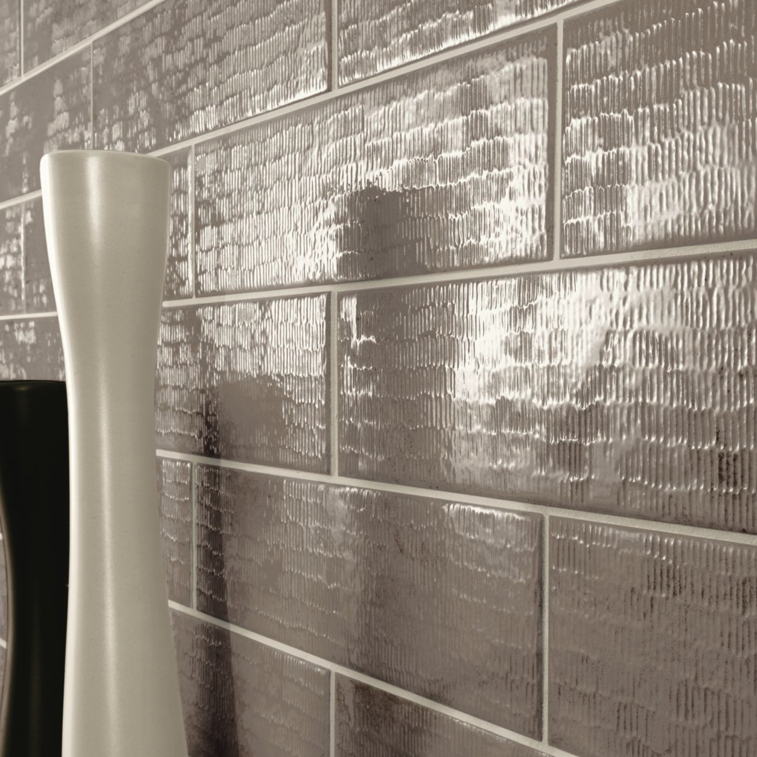 Close-up of a textured, glossy Inciso - Pebble silver subway tile wall with blurred ceramic vases in the foreground.