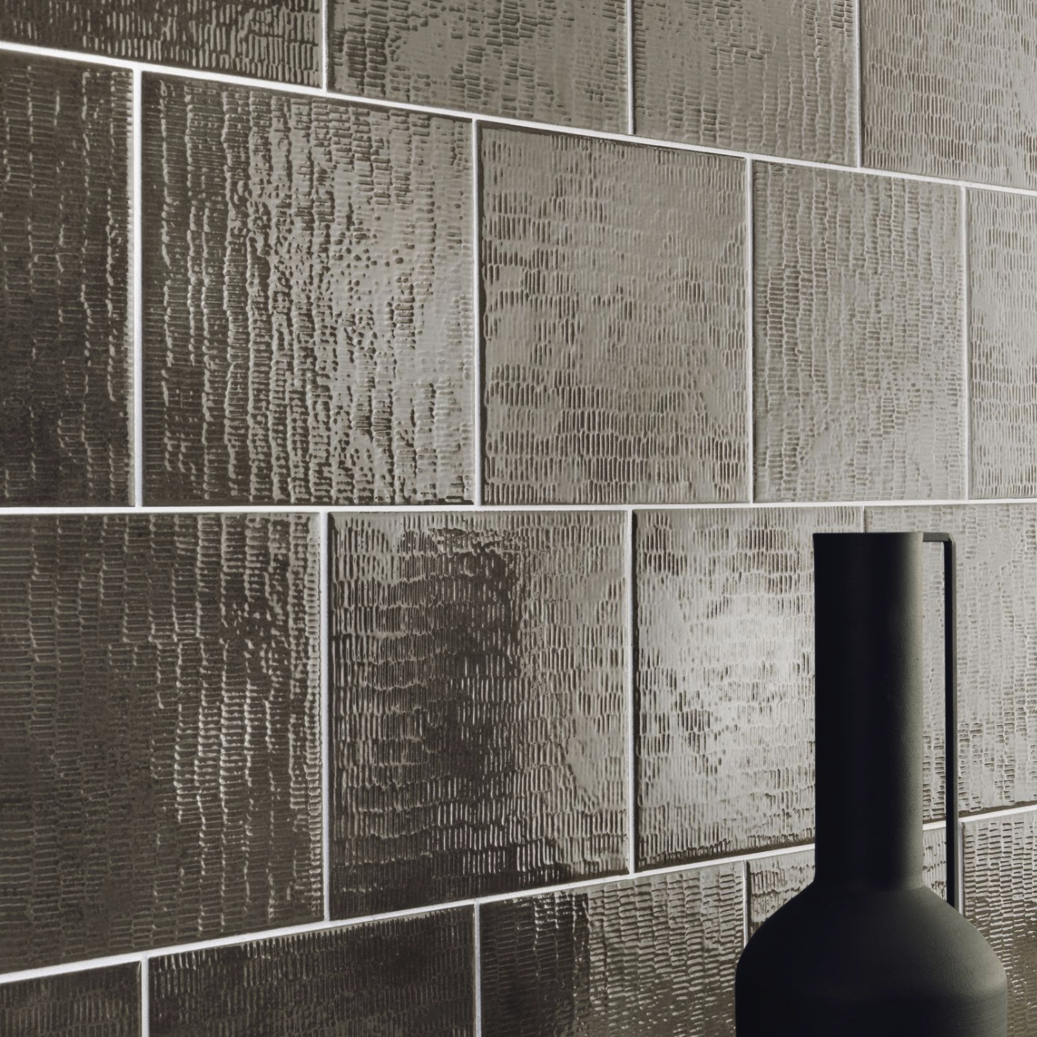 Close-up of Inciso - Porphyr tiles, showcasing their textured, glossy rectangular surfaces arranged in a staggered pattern on the wall. In the foreground, the top of a matte black handled vase is visible.