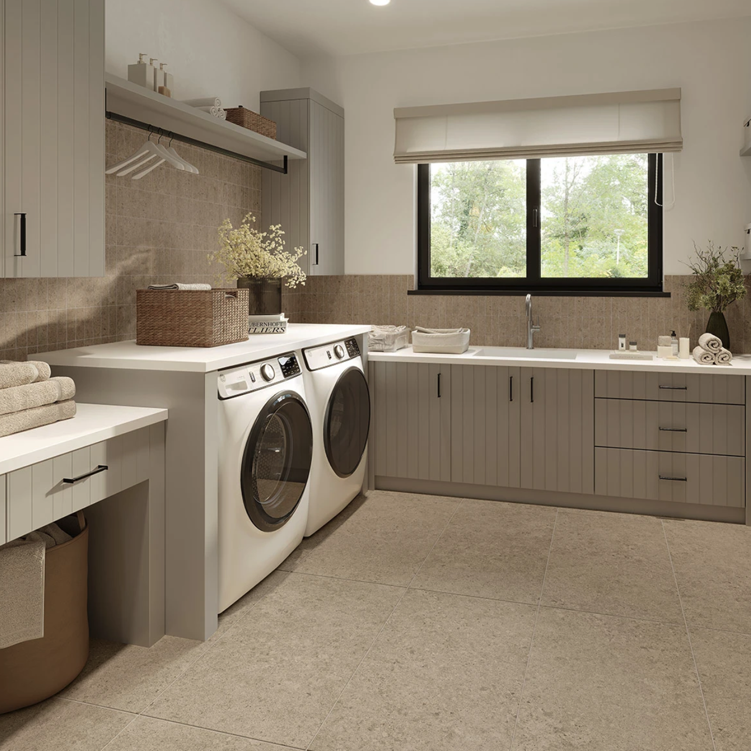 Modern laundry room featuring gray cabinets, a washer and dryer, countertop, sink, and a window offering views of lush trees. The space showcases clean organization with folded towels and plants as decor. The room is beautifully finished with Legacy Limestone - Clay, adding a refined touch to the overall design.