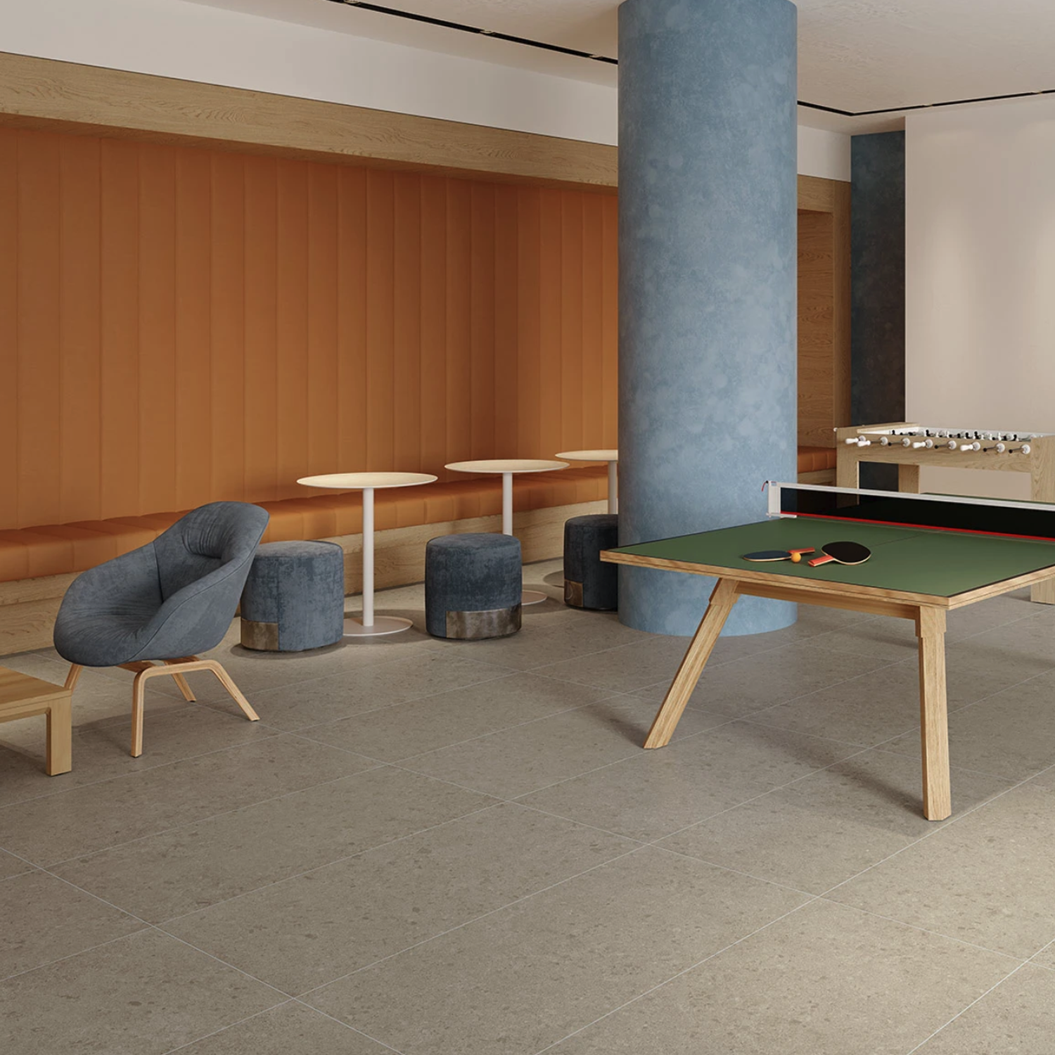 Modern lounge featuring Legacy Limestone - Harbor flooring, a ping pong table with two paddles and a ball, blue poufs, small white tables, a gray armchair, foosball table, orange wall panel, and a large blue pillar.