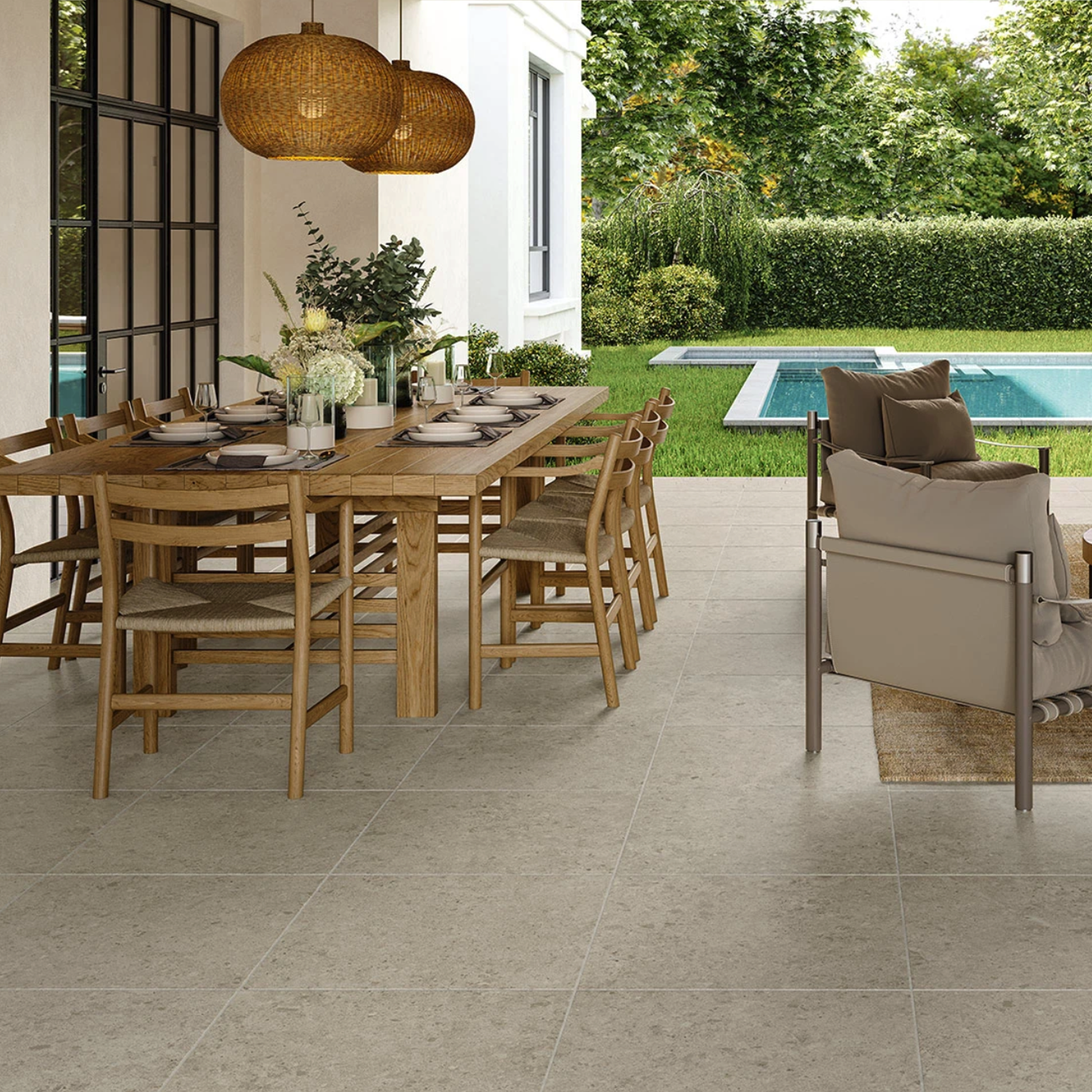 A spacious patio features the elegant Legacy Limestone - Silver flooring, complemented by a large wooden dining table, chairs, cushioned seating, pendant lights, potted plants, and a swimming pool set in a lush green backyard.