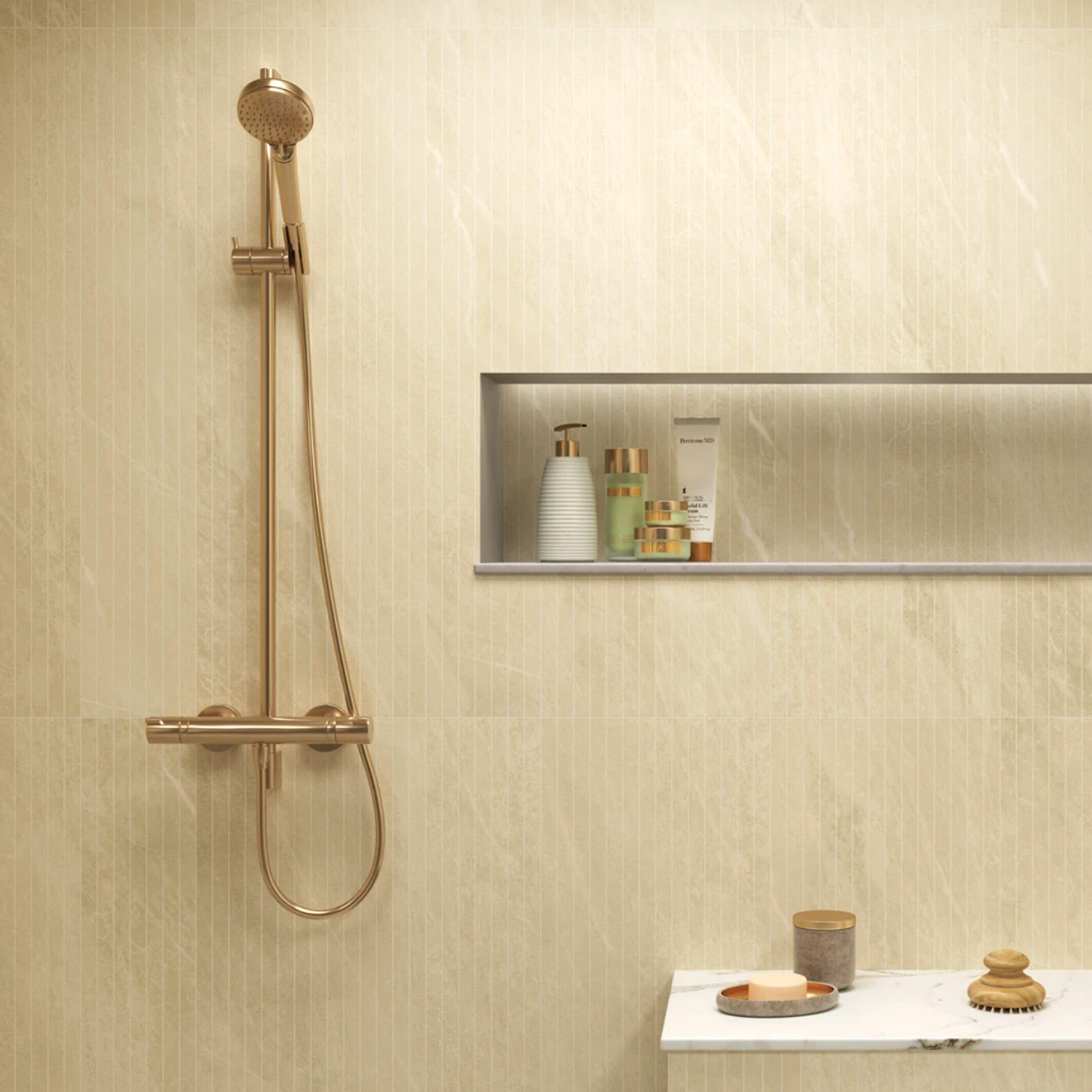 A modern bathroom featuring Legacy Marblestone - Sand tiles, complemented by a gold shower fixture, a recessed shelf with toiletries, and a marble counter adorned with a cup, soap, and brush.