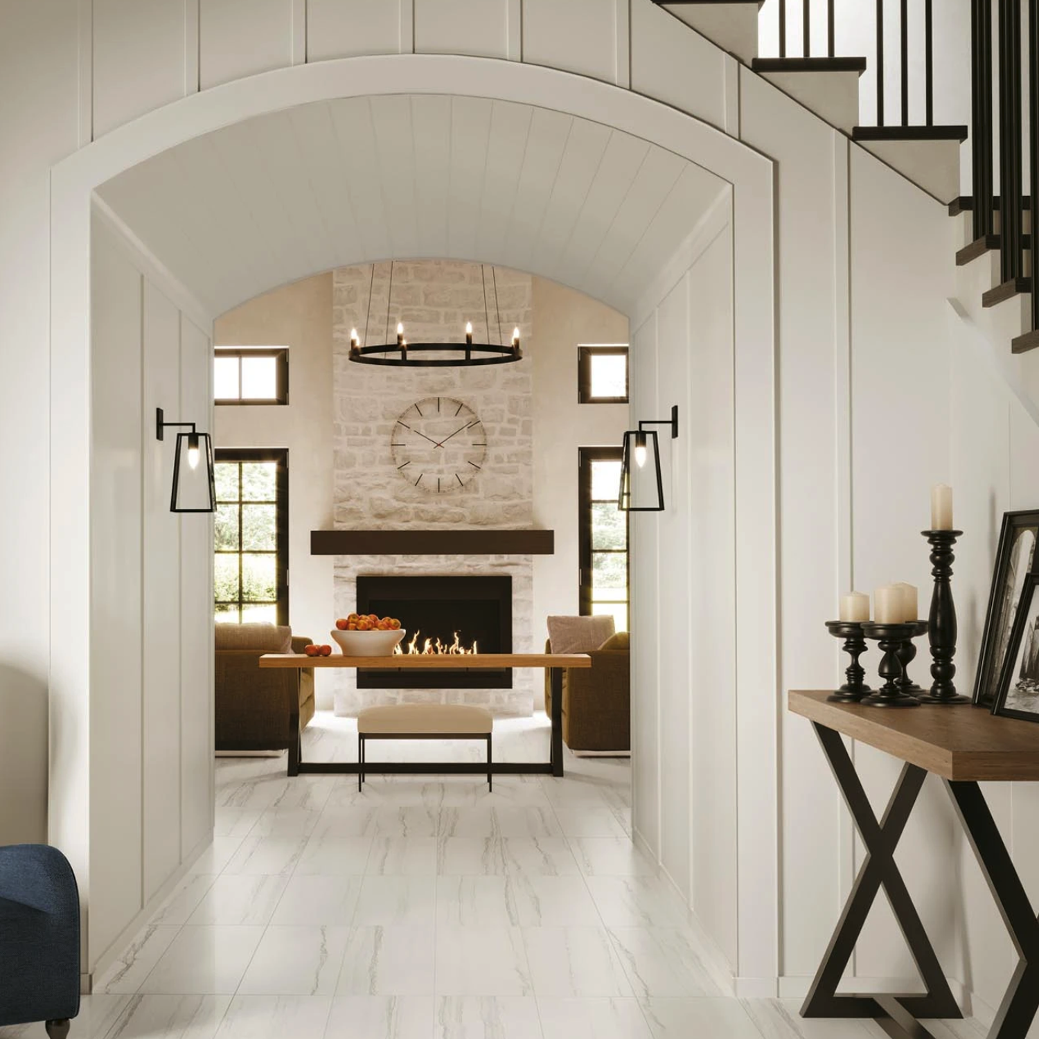 A modern entryway leads to a cozy living room featuring the Liberty - New England stone fireplace, a large clock, and brown sofas. Black sconces, a chandelier, and decorative candles provide elegant accents. White walls and light floors enhance the bright, airy atmosphere.