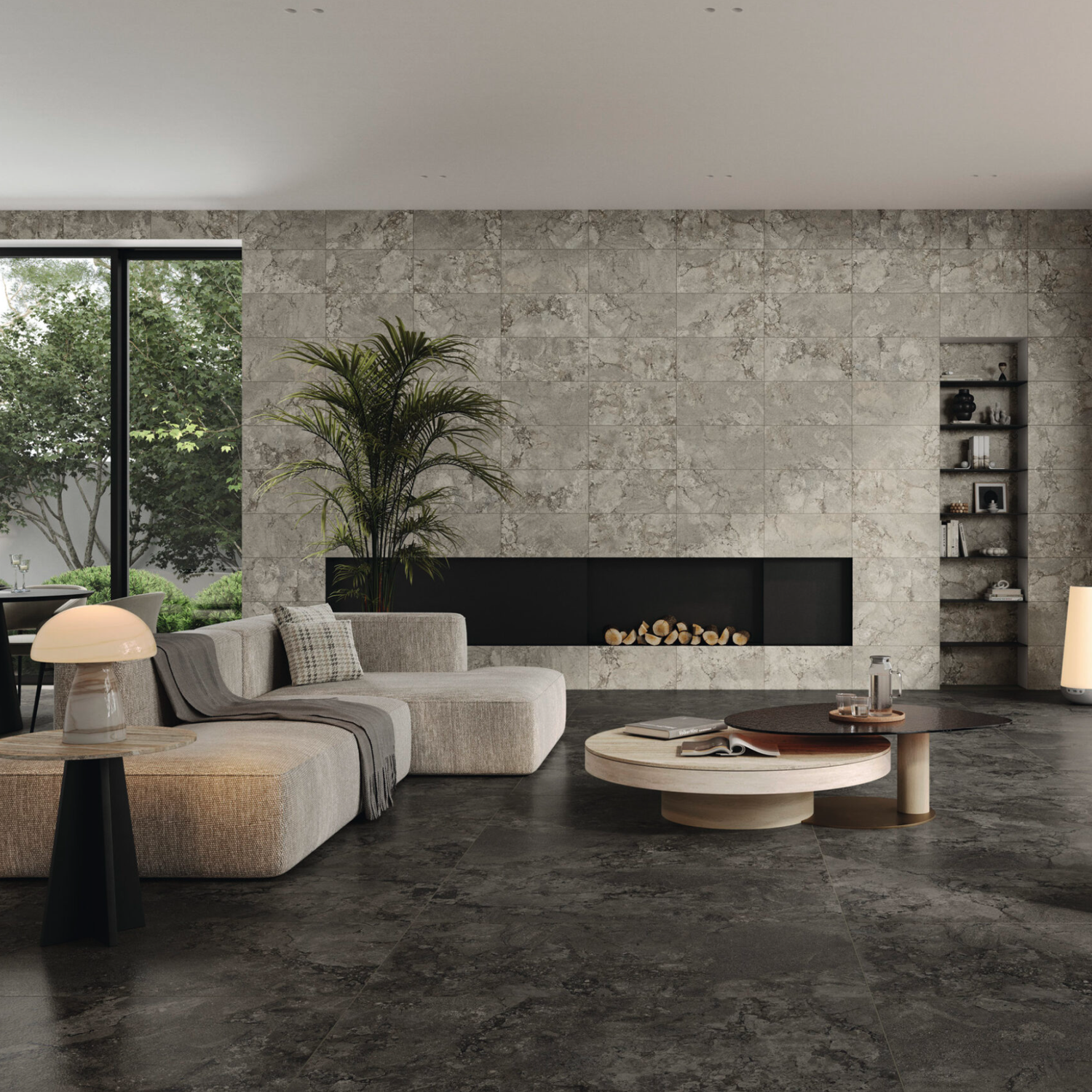Modern living room featuring Lythos - Coal gray stone walls and flooring, a beige sectional sofa, round coffee table, potted plant, built-in fireplace, shelves, and a large window with views of greenery outside. Minimalist decor in neutral tones.