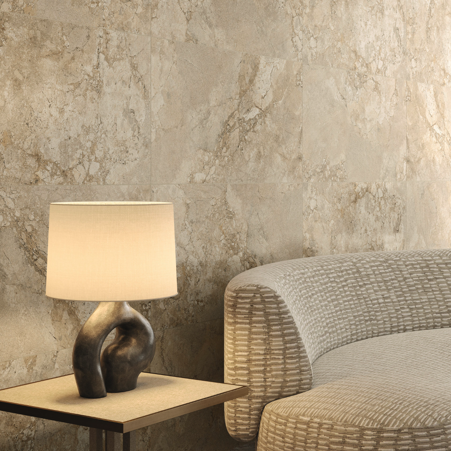 The Lythos - Sand table lamp, featuring a sculpted base, sits on a wooden table beside a textured, curved sofa. Behind them, the wall is adorned with beige, stone-patterned tiles. Warm lighting enhances the cozy and elegant atmosphere.