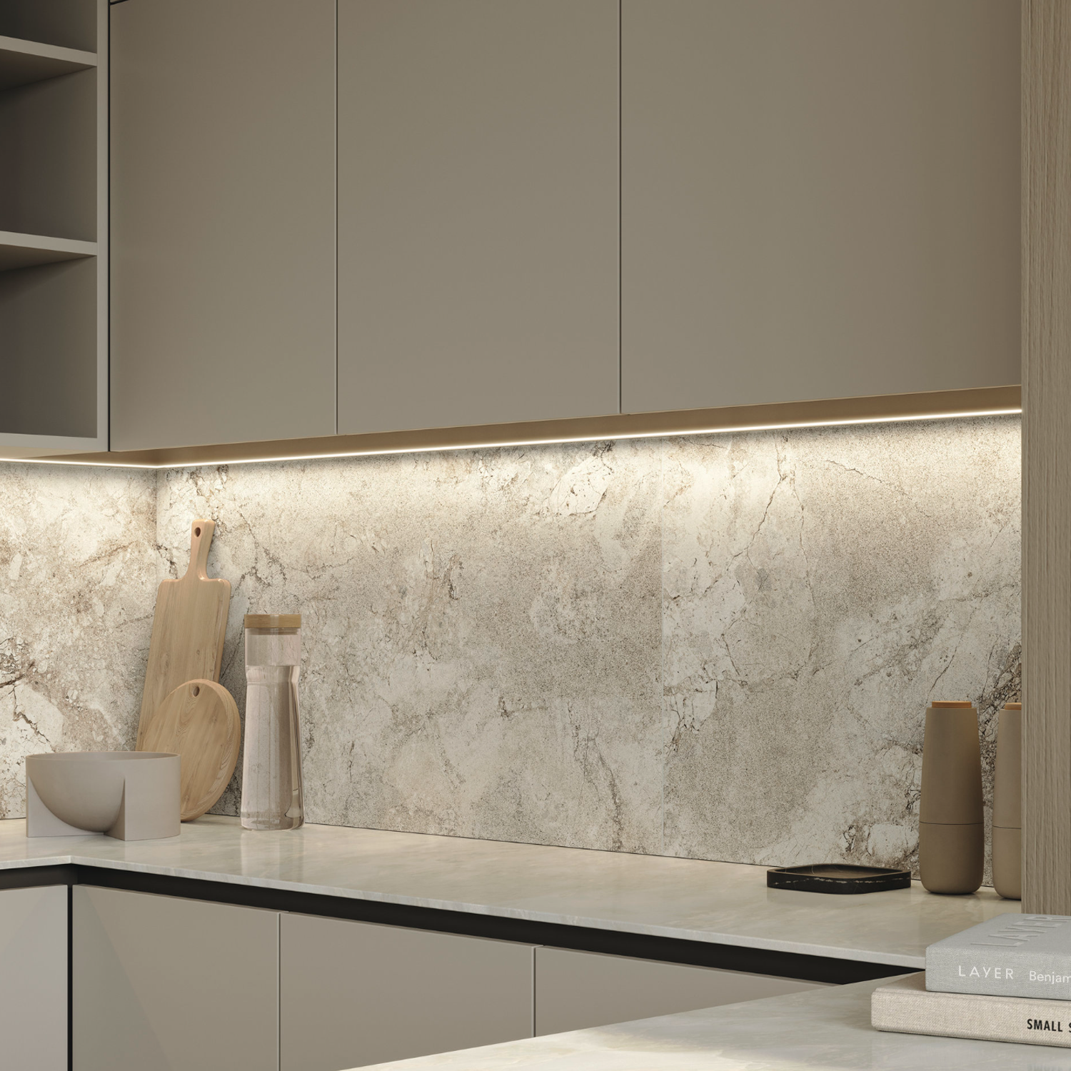 Modern kitchen featuring beige cabinets, a Lythos - Snow marble backsplash, under-cabinet lighting, and minimal decor such as wooden cutting boards, a bowl, a water bottle, and stacked books on the countertop.