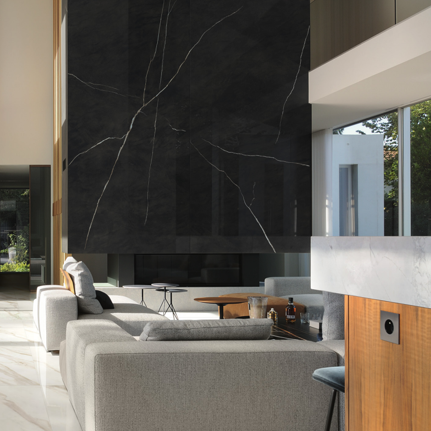 Modern living room featuring a large gray sectional sofa and low tables, highlighted by a striking Marvel Meraviglia - Black Origin marble accent wall with elegant white veining. Sunlight filters through tall windows, illuminating the light-colored floors and minimalist decor.