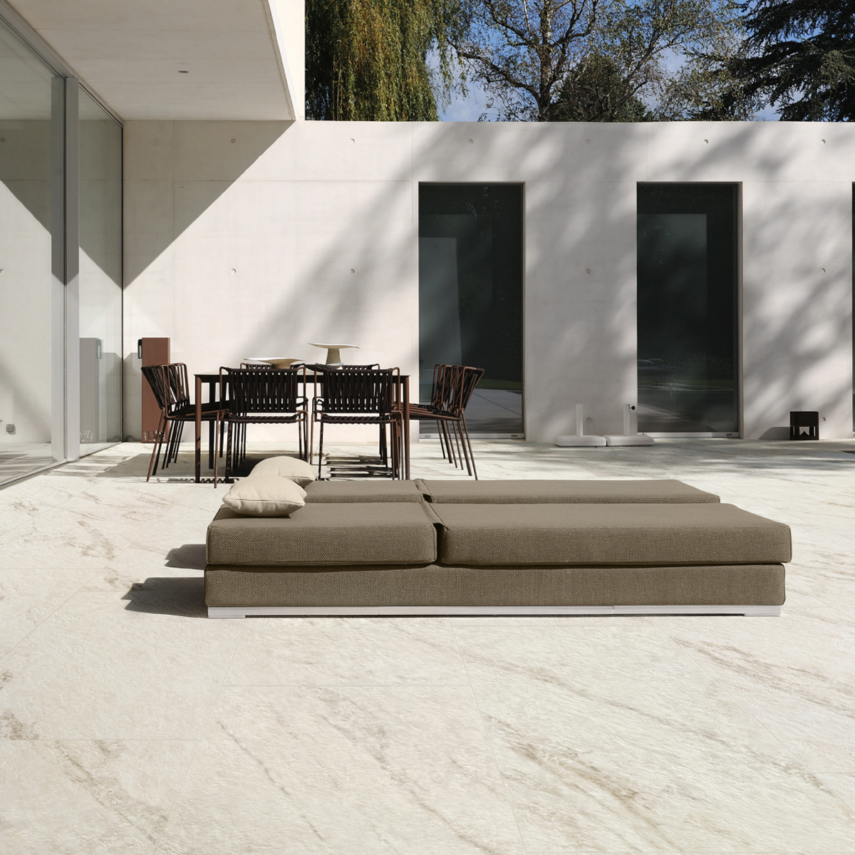 A modern patio featuring flooring in Marvel Meraviglia - Calacatta Bernini showcases a minimalist brown sofa with two beige pillows and a black dining set. The space is set against a white building with large glass windows and three tall dark doors, while trees and blue sky complete the scene in the background.