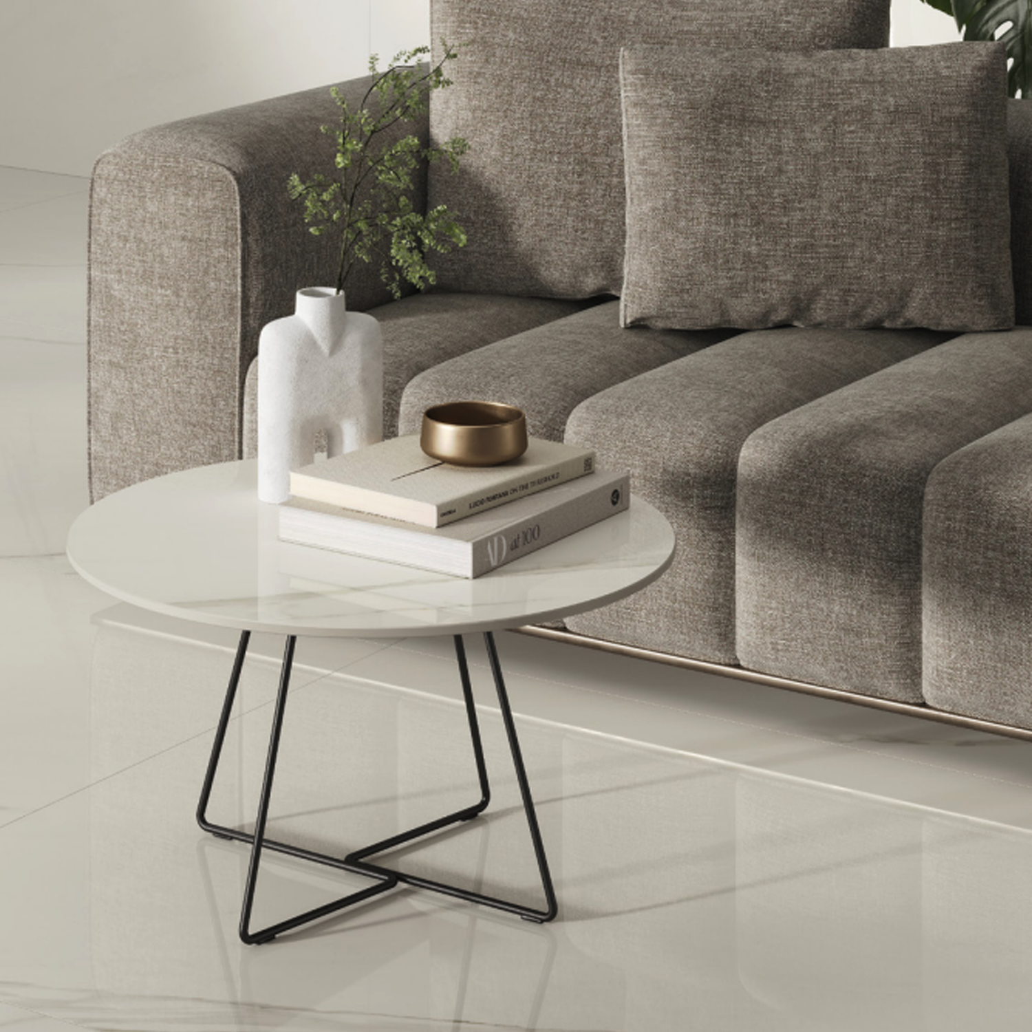A modern living room features a round white coffee table adorned with books, a white vase, a gold bowl, and green foliage in front of a textured gray sofa. The glossy white floors and walls are finished in Marvel Meraviglia - Calacatta Meraviglia for an elegant touch.