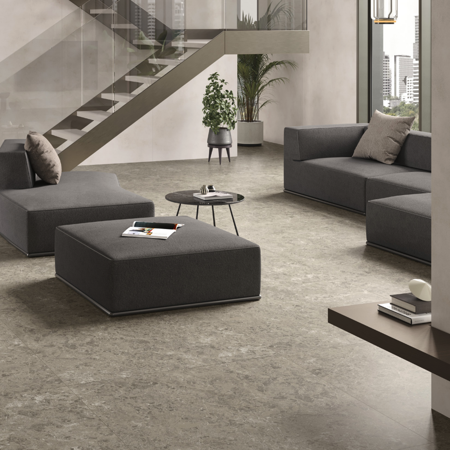 Modern living room featuring gray sofas and a low round table, complemented by indoor plants and a glass staircase. Floors are finished with Marvel Meraviglia - Grigio Elegante tiles, enhancing the contemporary look. Large windows reveal tall buildings outside, filling the space with natural light.