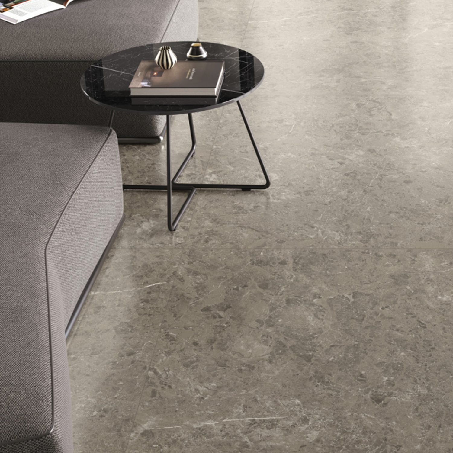A round black marble coffee table with a book and candle sits beside a modern gray sectional sofa on a large floor featuring Marvel Meraviglia - Grigio Elegante tiles.