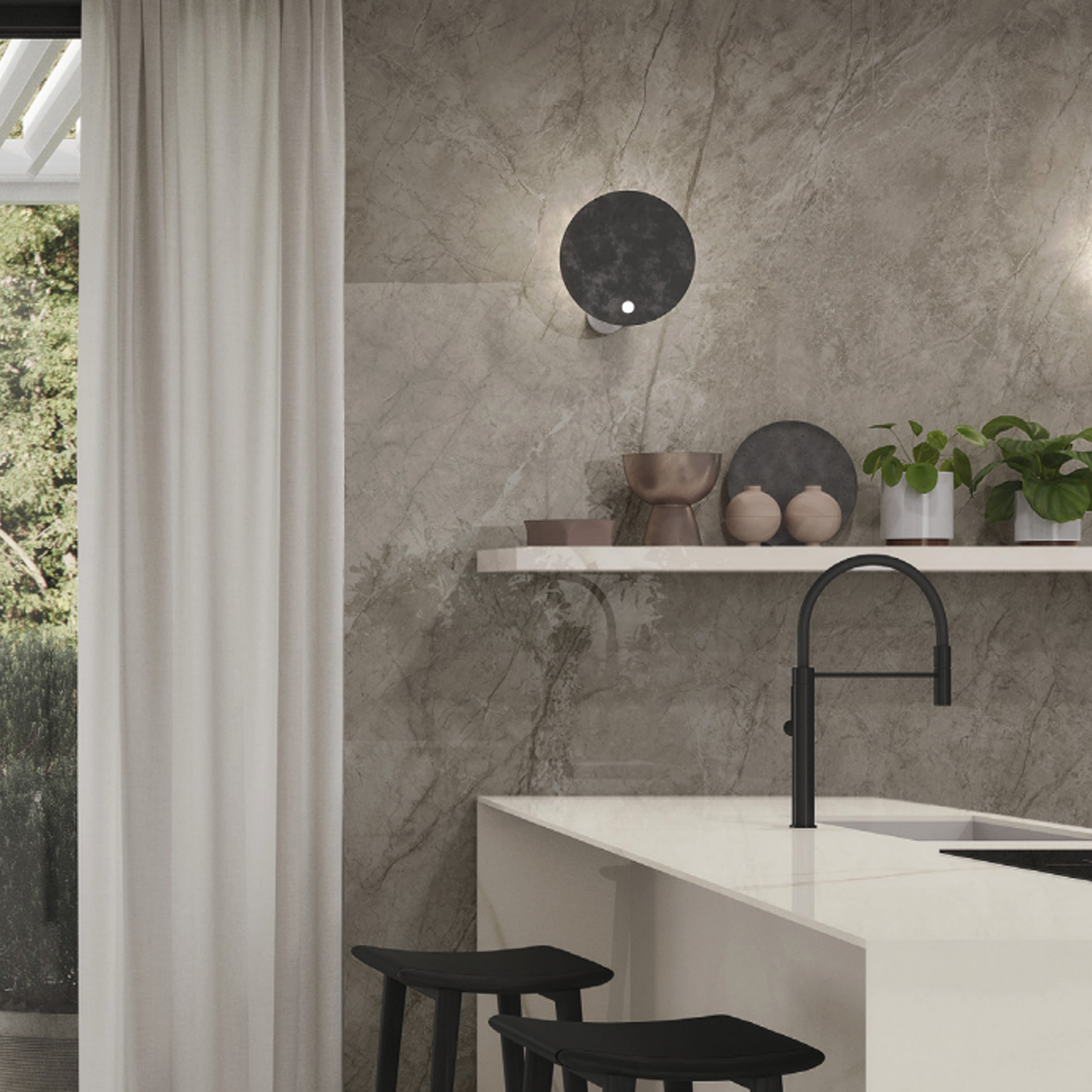 Modern kitchen featuring Marvel Meraviglia - Silver Majestic marble walls, a white island with a black faucet, two black stools, a floating shelf adorned with decor and plants, and a round black wall light. White curtains partially cover a large window overlooking lush greenery outside.
