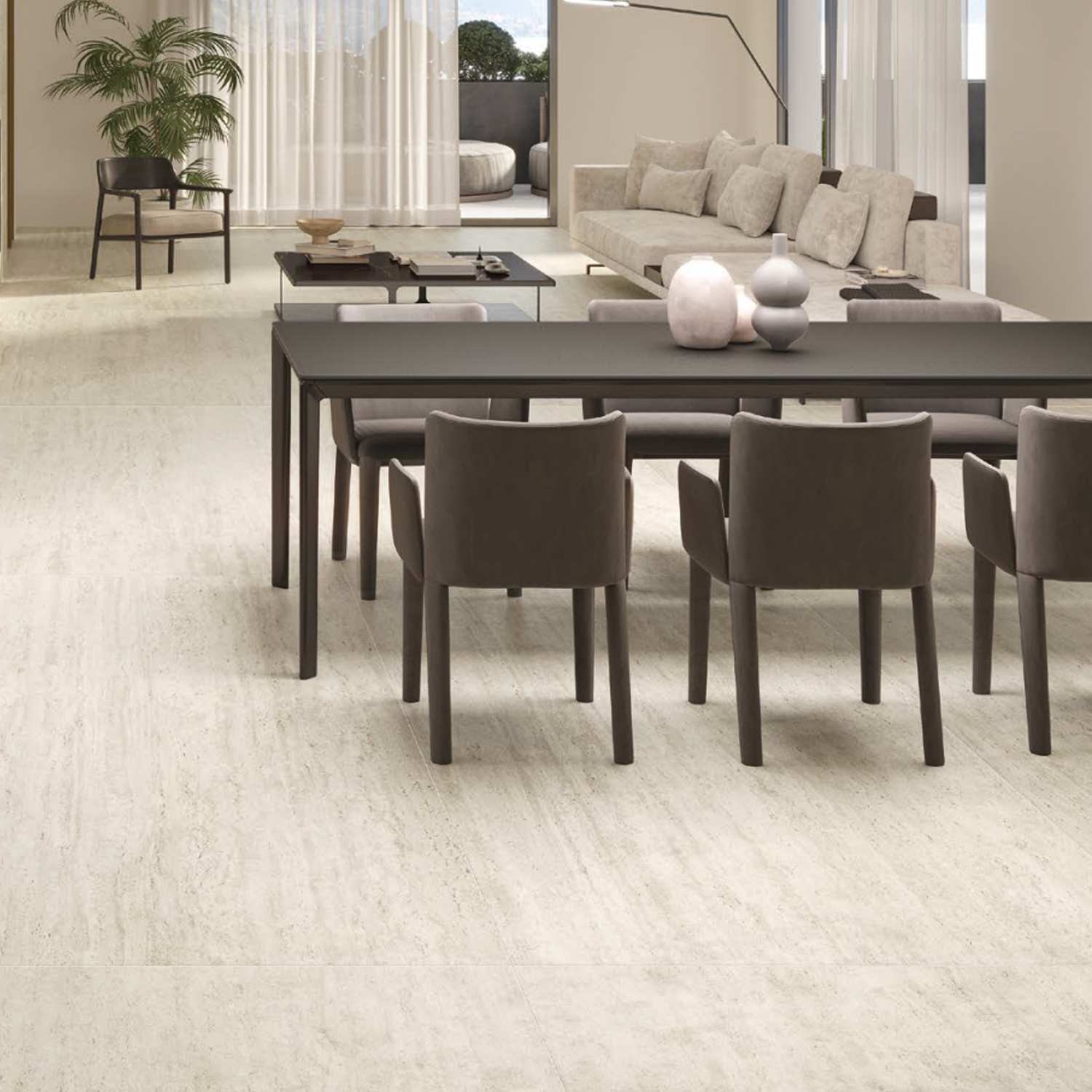 Modern dining room with a dark dining table, six brown chairs, Marvel Travertine - White Vein Cut tile floors, a light-colored sofa, and large windows with sheer curtains. A potted plant sits in the corner, and decorative objects are arranged on the table.