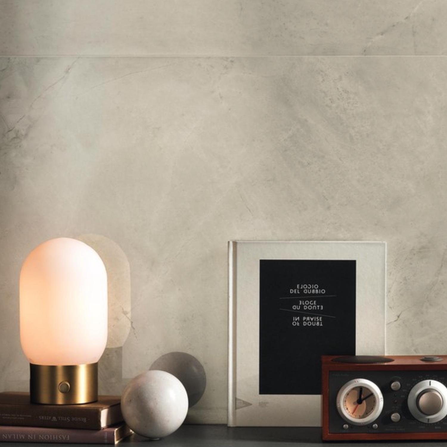 A modern table lamp with a frosted glass shade rests on a stack of books next to a framed print and a vintage-style radio, all beautifully arranged atop Marvel X - Calacatta Perla marble, set against a light gray stone wall backdrop.